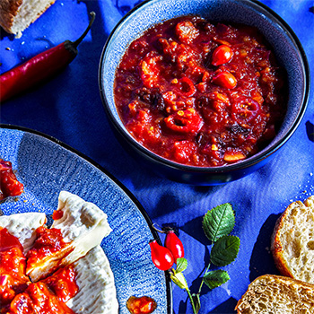 Foodfotografie eines Hagebutten-Chili-Dip in kleiner Schale, daneben ein Teller mit Camembert, auf den der Dip mit einem Teelöffel aufgetragen wurde; im Bild außerdem Baguette mit aufgeschnittenen Scheiben sowie dekorativ platzierte Chilischote und Hagebutten.