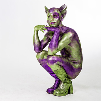 Glitzernder Mann in der Hocke, Arme auf den Knien abgestützt, Kopf ruht auf den Händen. Der Blick wirkt verträumt und geht aus dem Bild heraus. Ganzkörperaufnahme vor weißem Hintergrund.