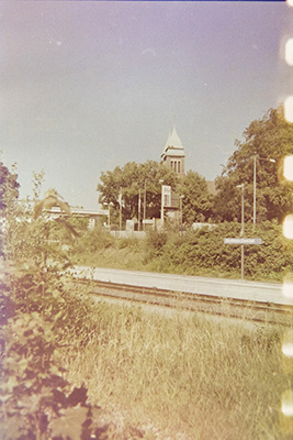 Bahn-Haltestelle mit Gleisen inmitten von Wiese und Büschen, im Hintergrund ist ein Kirchturm zu sehen.