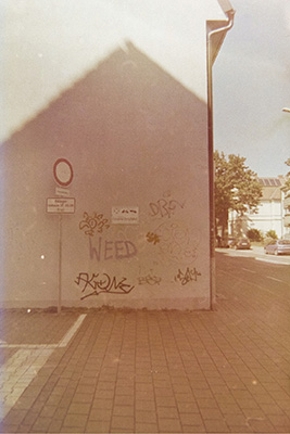 Ein helles Gebäude mit Graffiti-Tags, davor ein Verkehrsschild, Bürgersteig und ein Baum und Haus im Hintergrund.