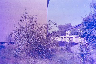 Bläuliche Aufnahme einer Hauswand mit kleinem Baum im Vordergrund. Im Hintergrund sind Garagen und ein Wohngebäude sichtbar.