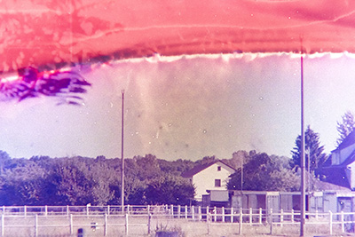 Querformat. Ähnliche Szenerie wie Bild 4, aber mit größerer Tiefe – viele Bäume im Hintergrund. Vom oberen Bildrand frisst sich ein rot-orangener Farbstreifen ins Bild.
