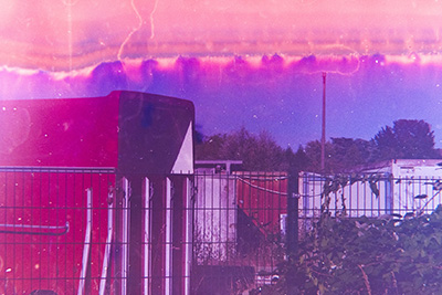 Querformat. Aufnahme mit Containern im Vordergrund, dahinter Bäume und blauer Himmel. Oben verläuft ein rot-orangener Streifen mit pinkem Saum.