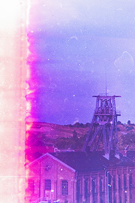Hochformat. Industriehalle mit einem Zechenturm. Im Hintergrund eine Landschaft mit Feldern und Bäumen. Am linken Rand eine orange-pinke Farbspur.