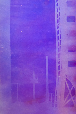 Hochformat. Rechts im Bild ein angeschnittener Metallmast. Im Hintergrund lassen sich schwach die Konturen einer Säule, Strommasten und eines Fernsehturms erkennen. Das gesamte Bild ist in Lila und Blau getaucht.