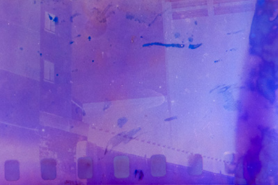 Querformat. Wieder sind unten die Löcher des Filmstreifens sichtbar. Inmitten der abstrakten lila-blauen Komposition lassen sich links Gebäudestrukturen mit Fenstern erahnen.