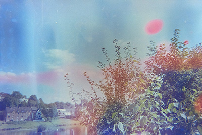 Querformat mit grünen Büschen im Vordergrund, Fluss und Wiesenlandschaft im Hintergrund. Blauer Himmel mit blassen Farbflecken. Eine horizontale rote Spur zieht sich durchs Bild, ergänzt durch einen roten runden Fleck rechts oben.